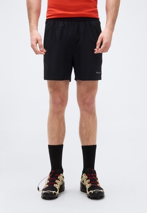 Mees, kes kannab musti sportlühikesi pükse, musti kesksäärega sokke ja musta-beezhika värviga maastikujooksu jalanõusid punaste paeltega, seisab valgel taustal.