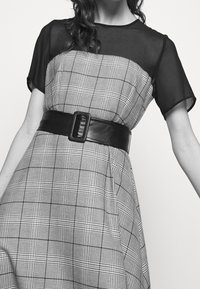 Robe noir et blanc avec un motif à carreaux ; manches courtes en tissu transparent ; large ceinture en cuir noir avec une boucle rectangulaire.