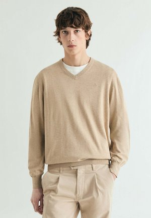 Joven con cabello castaño que lleva un suéter beige de cuello en V sobre una camisa blanca y pantalones beige, de pie contra un fondo claro y sencillo.