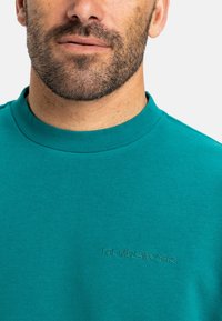 Teal bomulds sweatshirt med rund halsudskæring. Har broderet logo på brystet. Glat tekstur, kortærmede og afslappet pasform.