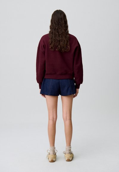 Sudadera de algodón burdeos combinada con pantalones cortos de mezclilla, zapatillas beige y calcetines claros. La tela parece suave y tiene un corte relajado.