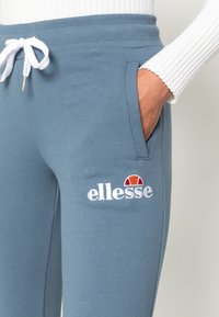 Blue sweatpants with white drawstring, featuring a side pocket and embroidered "ellesse" logo in white with a red and orange accent above it.