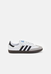 Zapatilla de cuero blanca con rayas negras, puntera de ante gris y suela de goma marrón. Cuenta con un logo azul en la lengüeta y texto dorado de la marca.