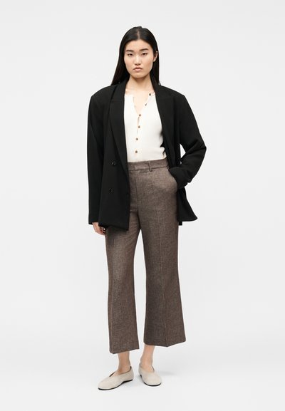 Femme debout portant un blazer noir oversize, un haut blanc boutonné, un pantalon large marron et des chaussures plates beige clair sur fond blanc uni.