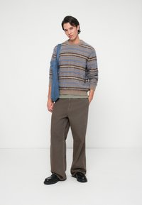 Sweater in soft, striped fabric with brown, blue, and beige hues; loose, long sleeves. Loose brown pants; black shoes; denim tote bag.