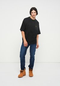 Svart oversized t-skjorte med en subtil grønn logo, kombinert med blå denimjeans og brune snølåsstøvler, på en ensfarget hvit bakgrunn.