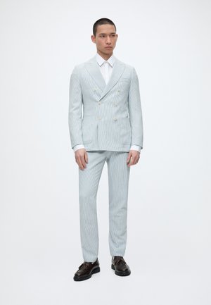 Mann trägt hellblauen zweireihigen Anzug mit weißem Hemd und dunkelbraunen Schnürschuhen, steht vor einem schlichten weißen Hintergrund.