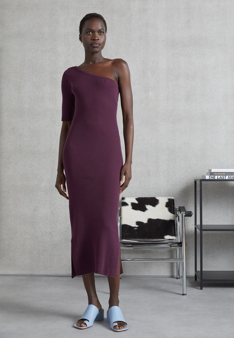 Vestido de malha canelada em bordô com design assimétrico de um ombro, até ao joelho, apresentando fendas laterais. Combinado com sapatos de salto aberto em azul claro.