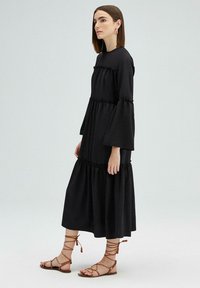 Zwarte maxi-jurk met gerimpelde lagen, lange klokmouwen en een ronde halslijn. Gecombineerd met bruine sandalen met banden tegen een lichte achtergrond.