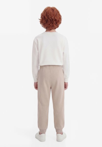 Barn med krøllete rødt hår står vendt bort, iført en hvit langermet skjorte, beige bukser med elastisk midje og hvite joggesko.