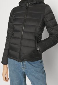 Personne portant une veste matelassée noire à capuche avec une fermeture éclair à l'avant et un jean bleu, une main dans la poche et l'autre détendue à côté.