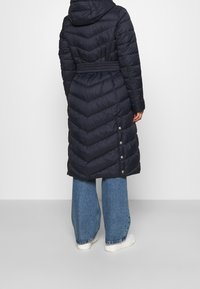 Marineblauer Steppmantel mit Kapuze, Chevron-Steppung, tailliertem Gürtel und Druckknöpfen an der Seite. Kombiniert mit weit geschnittenen Jeans und weißen Schuhen.