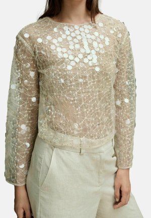 Femme portant un haut transparent beige à manches longues orné de sequins blancs circulaires et de motifs complexes en fil, associé à un pantalon beige clair.