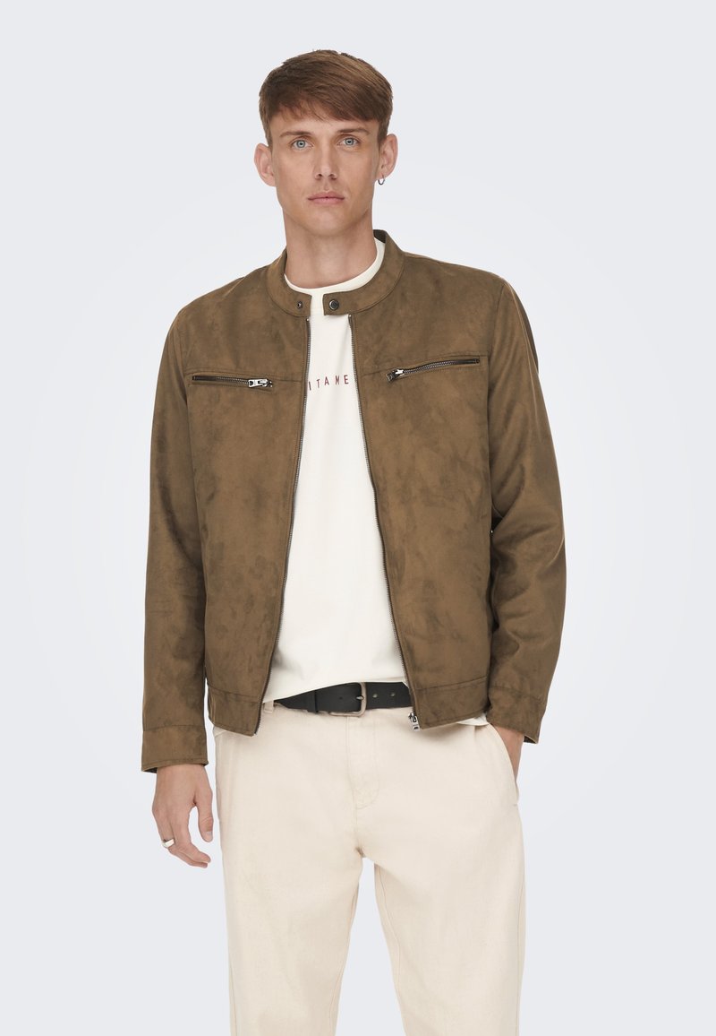 Jachetă bomber din suede maro, cu guler tip snap, două buzunare cu fermoar pe piept și o textură netedă, purtată peste un tricou de culoare deschisă și pantaloni crem.