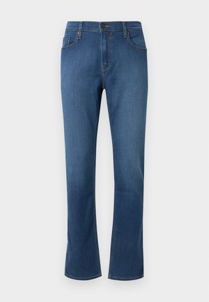 Mann steht, trägt ein schlichtes marineblaues T-Shirt, blaue Jeans und hellbeige Sneakers vor weißem Hintergrund.