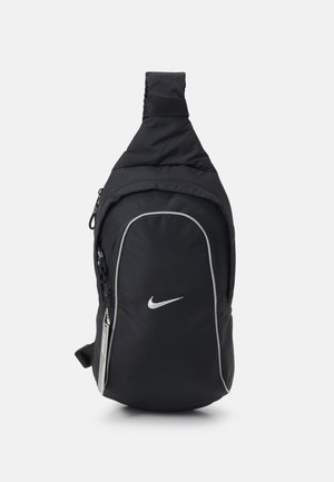 Sac à dos bandoulière noir Nike avec logo blanc, poche avant zippée et sangle réglable sur fond blanc uni.