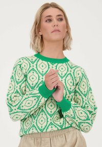 Groene en crèmekleurige trui met een geometrisch patroon, ribbelkraag en -manchetten in groen, en een zachte textuur.