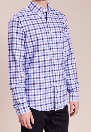 Hombre vistiendo una camisa de manga larga, abotonada, a cuadros azules y blancos, con pantalones negros, de pie frente a un fondo liso.