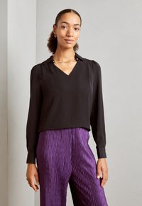 Blusa nera con scollo a V e maniche lunghe, abbinata a pantaloni viola texturizzati con un design plissettato. Dettaglio in catena d'oro al colletto.
