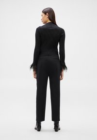 Sieviete stāv ar muguru pret kameru, valkājot melnas bikses ar augstu vidukli, ribētu garroku kreklu ar spalvotām aprocēm un melnus potīšu zābakus.