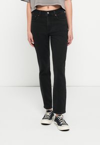 Jeans de mezclilla negra con un corte de pierna recta, que cuentan con cinco bolsillos y un cierre de botón. Combinados con zapatillas negras y blancas.