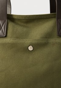 Τσάντα tote από καμβά σε ελιά πράσινο χρώμα με καφέ δερμάτινα λουριά, κλείσιμο με κουμπί και εμφανές καπιτονέ στα διακοσμητικά των λουριών.