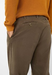 Bruine katoenen broek met een gladde textuur, voorzien van een knoopzak achter en zijzakken. De bovenste helft toont een oranje gebreide trui.