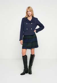 Μπλε μπουφάν με διπλό πέτο και ασημένια κουμπιά, συνδυασμένο με φούστα σε πράσινο και μπλε καρό. Το μοντέλο φοράει μαύρες μπότες μέχρι το γόνατο.