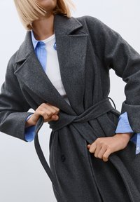 Gray wool coat with a belted waist, featuring a notch collar and two buttons. Underneath, a light blue collared shirt is visible.