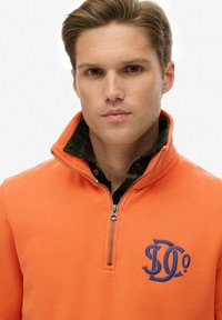 Orange zip-up fleece sweatshirt with blue logo detail. Features a standing collar and layered black and green checkered shirt underneath.