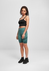 Fiatal nő áll, fekete crop topban, zöld, magas derekú kerékpáros shortban, fekete sportcipőben és ezüst láncnyakláncban, egy sima háttér előtt.