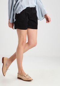 Femme portant un blouse bleu clair, un short noir ajusté et des mocassins en cuir beige, se tenant avec une jambe légèrement croisée devant.