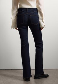 Jeans a zampa d'elefante blu scuro in denim, con vita alta, due tasche posteriori, cuciture arancioni a contrasto e una texture liscia.