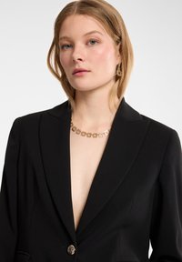 Blazer negro con un escote en V profundo, hombros estructurados y un detalle de botón dorado. Lleva un collar y pendientes con un patrón floral dorado.