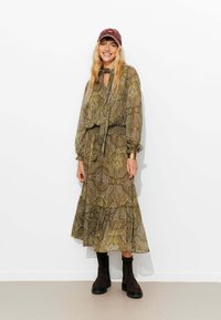 Robe longue et fluide de couleur verte avec des motifs paisley, associée à des bottines marron foncé et à une casquette bordeaux. Tissu transparent et détails texturés visibles.