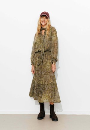 Robe longue et fluide de couleur verte avec des motifs paisley, associée à des bottines marron foncé et à une casquette bordeaux. Tissu transparent et détails texturés visibles.