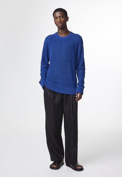 Königsblauer gestrickter Pullover mit strukturiertem Finish, Rundhalsausschnitt und gerippten Bündchen, kombiniert mit lockeren schwarzen Hosen und schwarzen Sandalen.