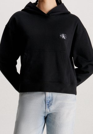 Osoba w czarnej krótkiej bluzie z kapturem z kieszenią z przodu i małym logo „cK”, w połączeniu z jasnoniebieskimi, wysokim stanem jeansami.