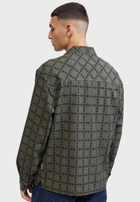 Cămașă cu imprimeu khaki, prezentând un model geometric în gri închis, mâneci lungi, guler clasic și hem curbat. Material texturat.