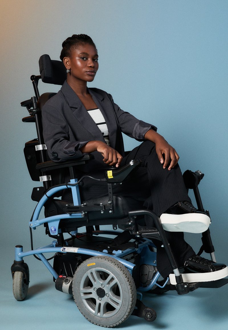 Elektrinė neįgaliųjų vežimėlis su mėlynu rėmu, juodu sėdyniu ir pilkiais ratais. Vartotojas vilki pilką švarką ir juodas kelnes, sėdi pasitikėdamas savimi.