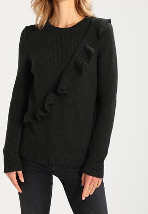 Woman wearing a black knitted sweater with a diagonal ruffle detail across the front and long sleeves, paired with black pants.