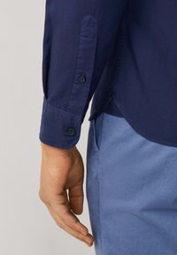 Marineblauwe shirtsmanchet met twee knopen en een tabdetail, met een gladde textuur. Lichtblauwe broek gedeeltelijk zichtbaar aan de onderkant rechts.