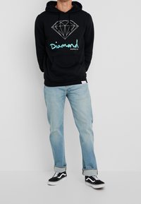 Svart hoodie med en geometrisk diamantgrafik och "Diamond Supply Co." i turkos, kombinerad med ljusblå jeans och svarta sneakers.