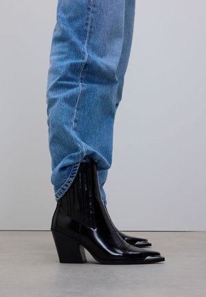 Persoon die lichte spijkerbroek draagt, in deze zijn glanzende zwarte cowboystijl enkellaarsjes met spitse neuzen en blokhakken gestoken, op een grijze vloer.