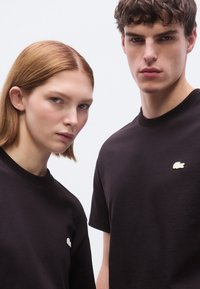 Δύο άτομα φορούν μαύρα t-shirts με στρογγυλή λαιμόκοψη, καθένα με μικρό λευκό λογότυπο Lacoste στο αριστερό στήθος. Απλός, χωρίς διακοσμήσεις σχεδιασμός.