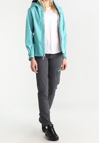 Lichtblauwe waterdichte jas met capuchon, grijze broek met een ritssluitingzak, en zwarte wandelschoenen met gestructureerde zolen.