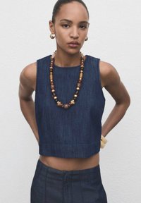 Denim crop top met een ronde halslijn, voorzien van een mouwloos ontwerp. Vergezeld van een ketting met gekleurde kralen en een armband van natuurlijk hout.