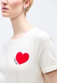 Γυναίκα που φοράει λευκό μπλουζάκι με κόκκινη καρδιά και τη λέξη "AMOUR" τυπωμένη κοντά στο στήθος, με κοντό ξανθό μαλλί και ασημένιο κρίκο σκουλαρίκι.