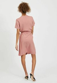 Růžové šaty s krátkým rukávem a stahovaním v pase, splývavý materiál, délka nad kolena, hladká textura a zakřivený lem. Nosí se s černými vysokými podpatky.