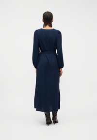 Γυναίκα με πλεξούδες, που φοράει μακρύ μπλε φόρεμα με φουσκωτά μακριά μανίκια και μαύρα μποτάκια με τακούνι, στέκεται μπροστά σε απλό λευκό φόντο.